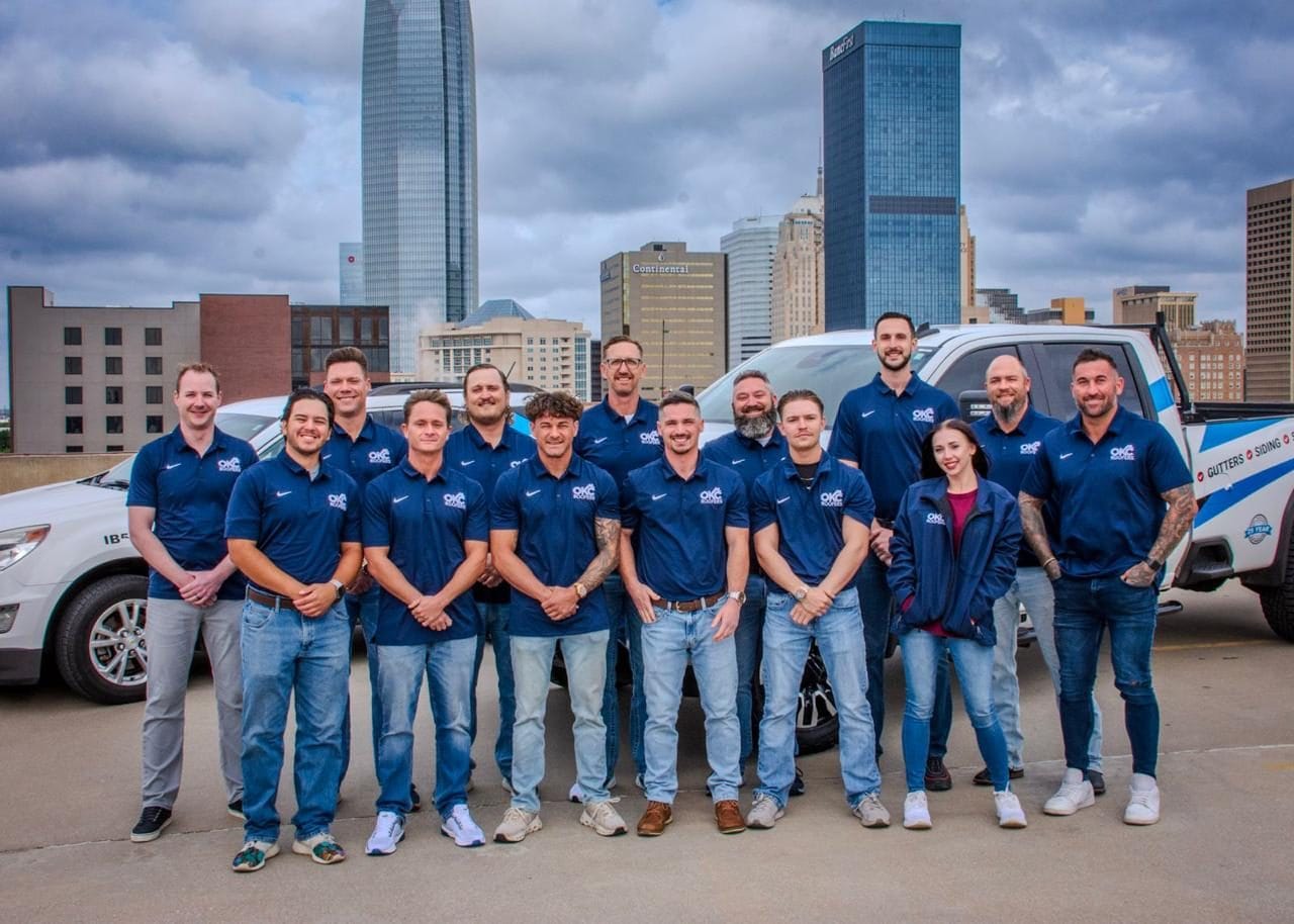 Full OKC Roofers company team of fourteen staff members in front of branded service trucks with the downtown Oklahoma City skyline including Devon Tower and BancFirst Tower